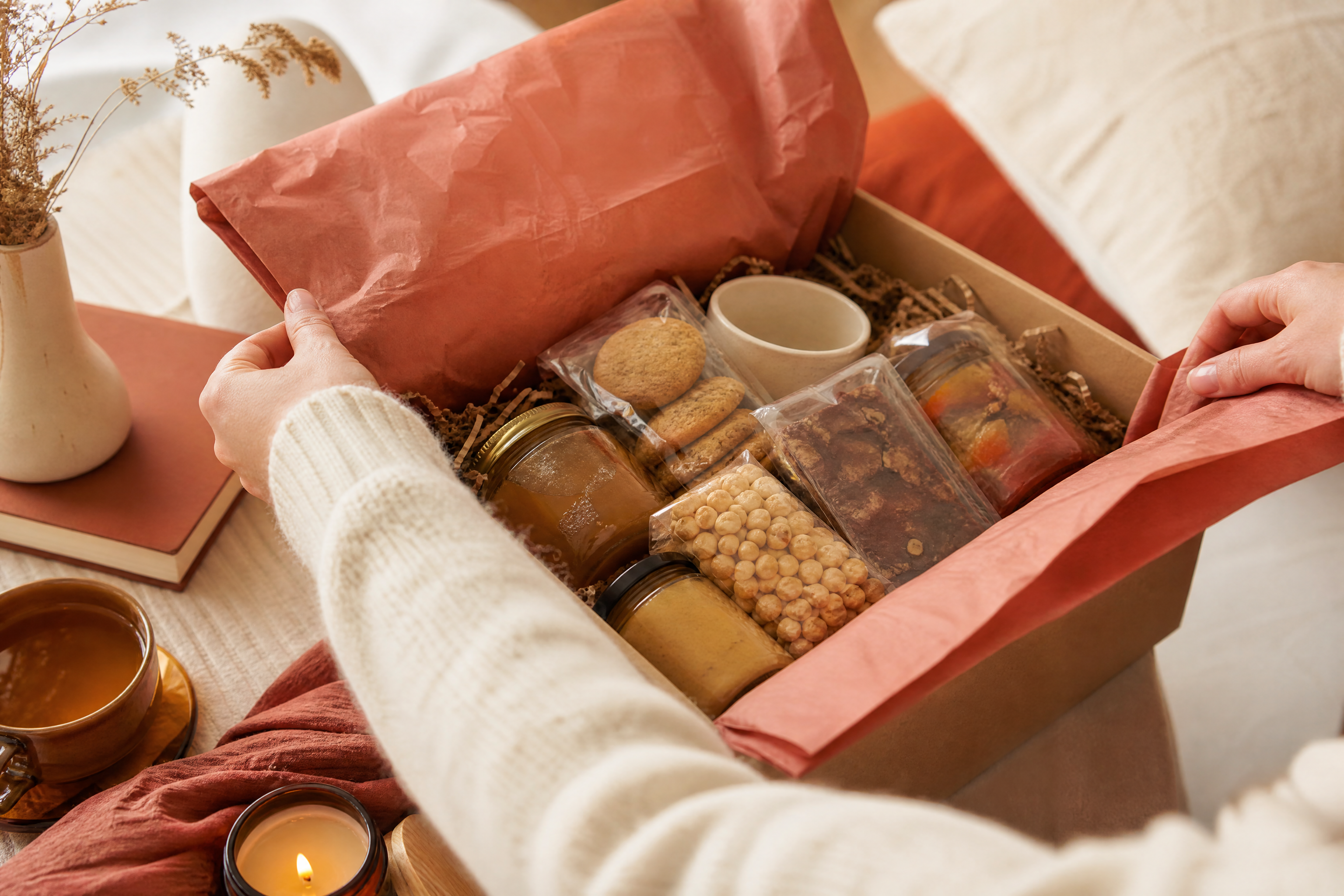 Hands opening a gift box of assorted packaged foods on a cozy table.